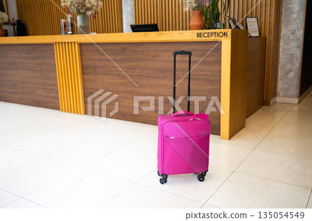 Pink suitcase standing in hotel lobby interior. Travel readiness, movement pause, and modern tourism lifestyle symbolizing transition, journey anticipation, and temporary accommodation experience Pink suitcase standing in hotel lobby interior. Travel readiness, movement pause, and modern tourism lifestyle symbolizing transition, journey anticipation, and temporary accommodation experience 135054549
