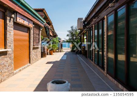 Narrow seaside alley between buildings leading toward blue sea. Architectural framing, travel anticipation, and calm coastal transitions guiding visitors toward leisure spaces. Narrow seaside alley between buildings leading toward blue sea. Architectural framing, travel anticipation, and calm coastal transitions guiding visitors toward leisure spaces. 135054595
