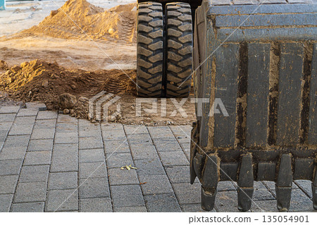 Construction site in which an excavator with lowered bucket to the surface of street tiles. Specialized machines for excavation, laying and replacement of pipes, underground utilities. Working place. Construction site in which an excavator with lowered bucket to the surface of street tiles. Specialized machines for excavation, laying and replacement of pipes, underground utilities. Working place. 135054901