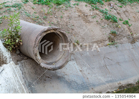 The end of old concrete pipe of large diameter protrudes from the ground above a ditch with a flowing small river with reinforced concreted banks. Road underground communications, symbol of road end 135054904