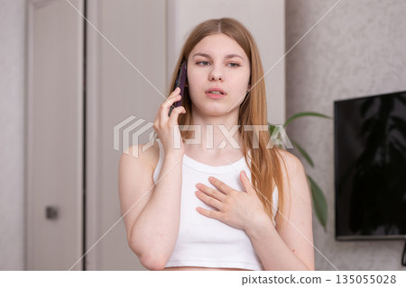 Young woman talking on the phone in a modern living room during the day while expressing concern or surprise Young woman talking on the phone in a modern living room during the day while expressing concern or surprise 135055028