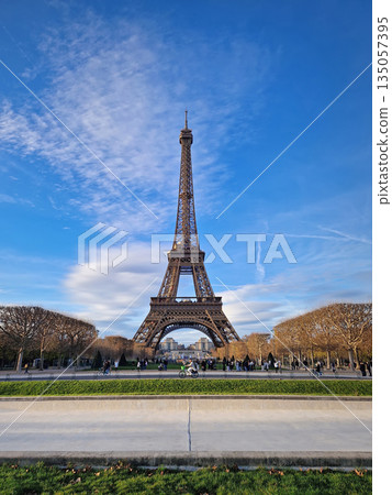 Paris, France - December 09, 2025: the majestic Eiffel Tower rising high into a bright blue sky filled with soft, wispy clouds. Crowds of people stroll through the green Champ de Mars below 135057395