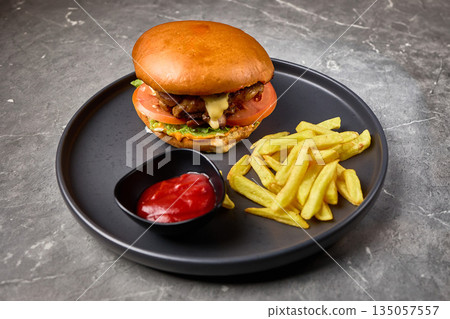 Delicious burger with tomato, lettuce, accompanied by fries and red sauce on black plate, fast food concept 135057557