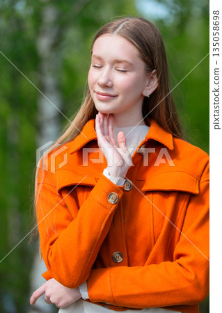Young woman in orange coat enjoying nature outdoors on a sunny day with trees in the background 135058698