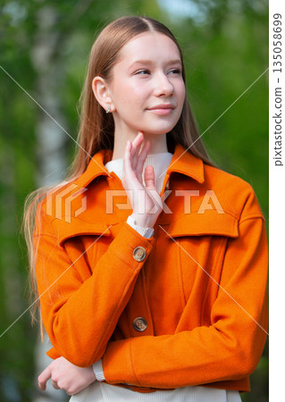 Young woman in orange jacket poses thoughtfully outdoors surrounded by greenery in early spring light Young woman in orange jacket poses thoughtfully outdoors surrounded by greenery in early spring light 135058699