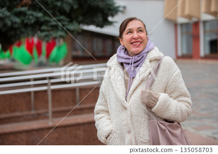 A middle-aged woman in a white fur coat in an urban setting. She smiles and does not look at the camera. 135058703