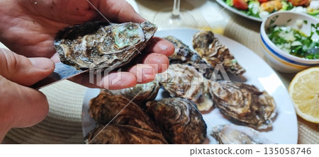 A close-up of hands opening fresh oysters with a knife over a plate. Concepts of fine dining, seafood delicacies, restaurant dining, home cooking, nutrition, and authentic gastronomic experiences. 135058746