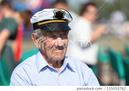 Elderly man in captain's hat enjoys sunny outdoor event with friends at a festive gathering 135058762