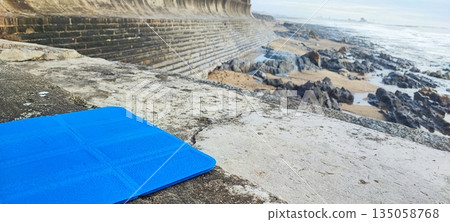 A brightly colored yoga or fitness mat resting on concrete steps by the ocean shore. The concept of mindfulness, outdoor exercise, healthy living, meditation and balance, stress relief, and self-care 135058768