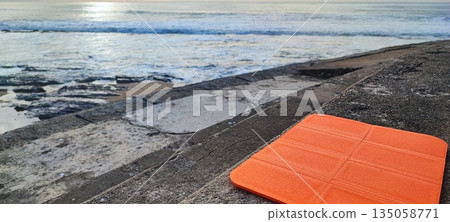 A brightly colored yoga or fitness mat resting on concrete steps by the ocean shore. The concept of mindfulness, outdoor exercise, healthy living, meditation and balance, stress relief, and self-care 135058771