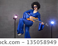 A girl in a black wig poses with a guitar in a vintage photo shoot in the style of the eighties. 135058949