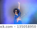 Girl comic pop star posing in black wig with toy guitar on blue background. Rock style of seventies 135058950