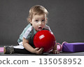 A young child is playing with a red ball. The child is sitting on the floor and holding the ball in his hand 135058952