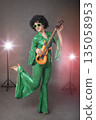 Fun eighties style. Girl comic pop star posing in black wig with toy guitar on gray background. 135058953