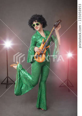 Fun eighties style. Girl comic pop star posing in black wig with toy guitar on gray background. 135058953