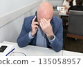 A tired businessman is talking on his smartphone and he covered his face with his hands. 135058957