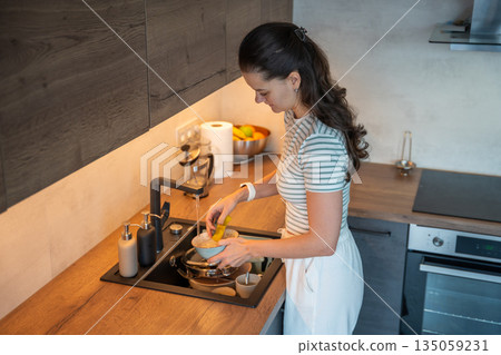Woman washing dishes at kitchen sink after cooking at home. Everyday household cleaning task and hygiene routine in real life interior 135059231