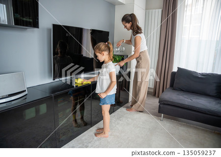 Mother and daughter cleaning TV screen in living room at home. Everyday household routine and family cooperation in real life interior Mother and daughter cleaning TV screen in living room at home. Everyday household routine and family cooperation in real life interior 135059237