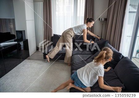 Mother and daughter arrange sofa cushions together in a living room at home. Real life family scene showing everyday routine, cooperation, shared responsibility and domestic organization. 135059239