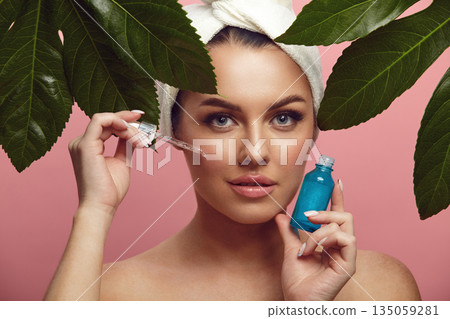 Beautiful young woman in head towel holding facial serum stands among tropical leaves over pink isolated background. Advertisement of beauty product. Beautiful young woman in head towel holding facial serum stands among tropical leaves over pink isolated background. Advertisement of beauty product. 135059281