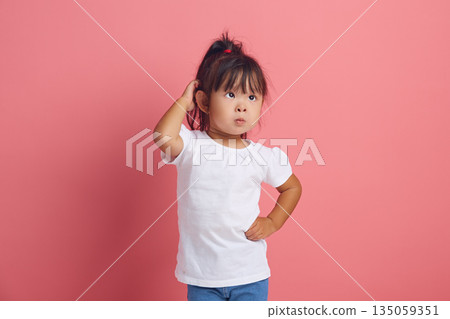 Confused little child scratches her head with thoughtful look has a puzzled expression dressed in neutral white t shirt looks with puzzlement aside, poses against pink background. Confused little child scratches her head with thoughtful look has a puzzled expression dressed in neutral white t shirt looks with puzzlement aside, poses against pink background. 135059351