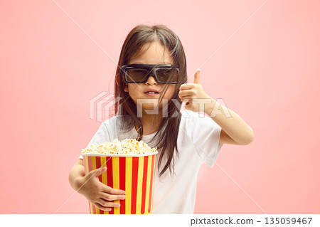 Enthusiastic little kid girl Movie Fan with Popcorn Approves of Film Time standing on Pink Background Enthusiastic little kid girl Movie Fan with Popcorn Approves of Film Time standing on Pink Background 135059467
