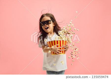 Shocked Surprised Little Kid Girl 5-6 years Old in 3D Glasses Watching Movie Film in Cinema, Holding Bucket of Popcorn Flying to the Different Sides, Standing on Pastel Pink Isolated Background. Shocked Surprised Little Kid Girl 5-6 years Old in 3D Glasses Watching Movie Film in Cinema, Holding Bucket of Popcorn Flying to the Different Sides, Standing on Pastel Pink Isolated Background. 135059474