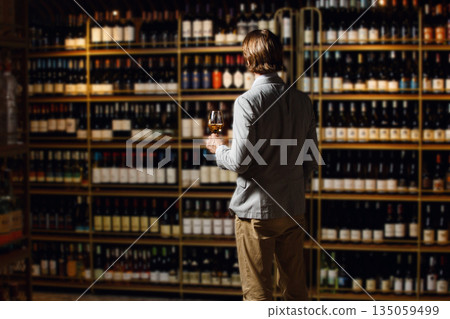 Back portrait of experienced and skilled sommelier holding wine glass and looking to cellar with wooden bottle filled shelf. Male bartender studies different varieties of wine by region. 135059499