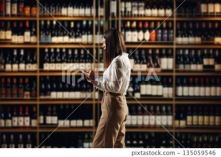 Female expert sommelier locating in winery sphere analyses various varieties of red and white wine, located in wine cellar, making notes in tablet. Female expert sommelier locating in winery sphere analyses various varieties of red and white wine, located in wine cellar, making notes in tablet. 135059546