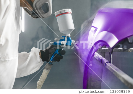Car paint worker spraying purple paint to car body element using spray gun in vehicle workshop chamber. Complex restoration of automobile body. 135059559