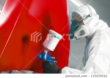Worker in protective mask and overall spraying base coat of paint on car body elements in painted chamber using professional airbrush. 135059582