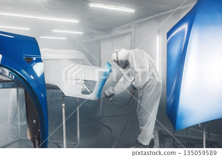 Car paint worker spraying blue paint to car body element using spray gun in vehicle workshop chamber. Complex restoration of automobile body.  135059589