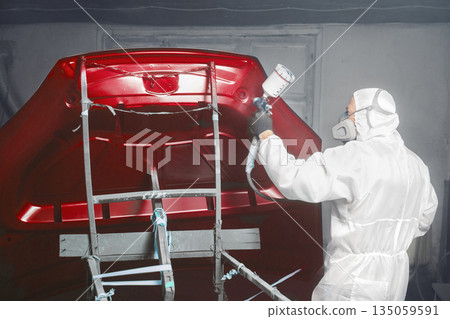 Professional spray painter in protective mask and clothes spraying car hood of vehicle part to red color using spray gun in paint chamber.  135059591