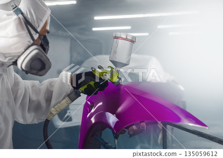 Car paint worker spraying purple paint to car body element using spray gun in vehicle workshop chamber. Complex restoration of automobile body. Car paint worker spraying purple paint to car body element using spray gun in vehicle workshop chamber. Complex restoration of automobile body. 135059612