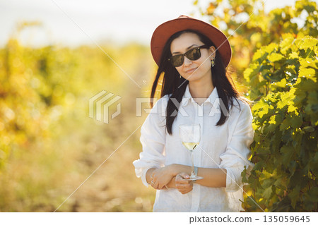 Woman in Hat and Sunglasses Standing in Field of Lush Foliage of Grapes 135059645