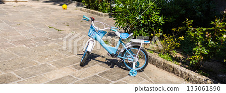Blue kids bicycle with training wheels on sunny pavement, banner with copy space. Childhood fun, outdoor learning, and the joy of early cycling experiences in a quiet garden path. 135061890