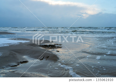 Winter evening on the Baltic Sea coast with icy waves, wet sand, foam patterns and dramatic sky Winter evening on the Baltic Sea coast with icy waves, wet sand, foam patterns and dramatic sky 135061910