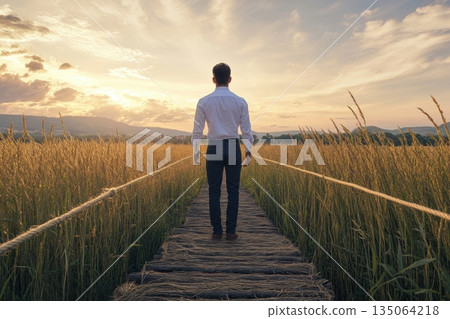 Man standing on wooden path in golden field during sunset with vibrant sky and distant mountains 135064218