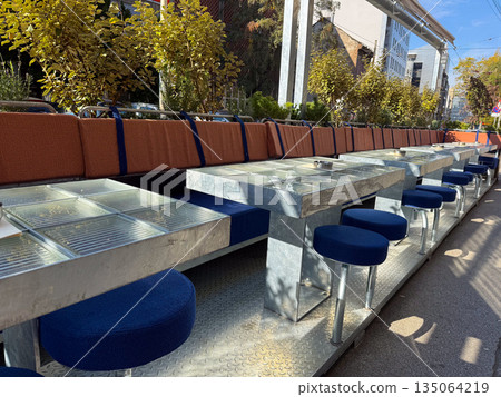 Outdoor cafe seating area with metal tables and blue stools on a sunny city street. Modern urban design, daylight shadows, and warm autumn atmosphere. Outdoor cafe seating area with metal tables and blue stools on a sunny city street. Modern urban design, daylight shadows, and warm autumn atmosphere. 135064219