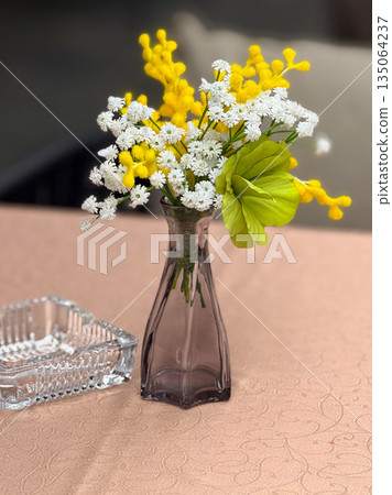 Bright yellow and white flowers in a glass vase on a cafe table. Elegance, simplicity, and cozy atmosphere in a calm urban setting. Bright yellow and white flowers in a glass vase on a cafe table. Elegance, simplicity, and cozy atmosphere in a calm urban setting. 135064237