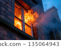 Flames erupt from an upper floor window of a building during a fire incident at dusk 135064943
