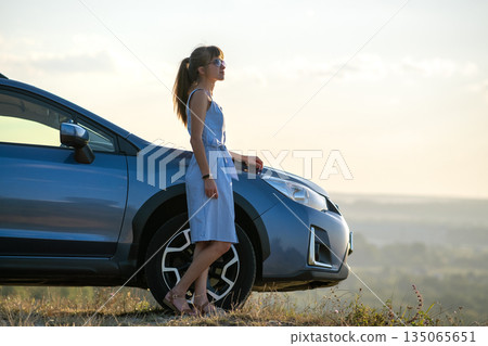 Happy young woman in blue dress standing near her vehicle looking at sunset view of summer nature. Travelling and vacation concept. 135065651