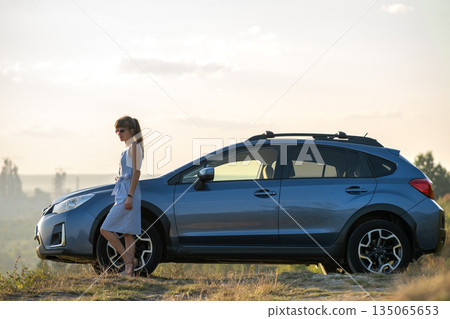 Happy young woman driver in blue dress enjoying warm summer evening standing beside her car. Travelling and vacation concept. 135065653