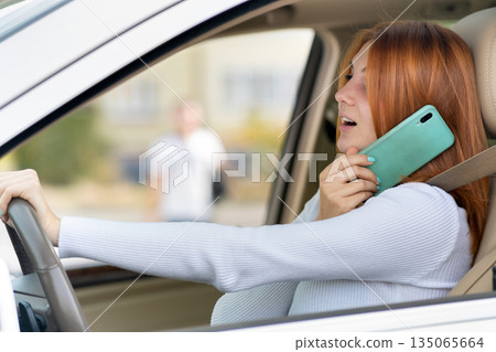 Happy redhead girl talking on her mobile phone behind the wheel driving a car. 135065664