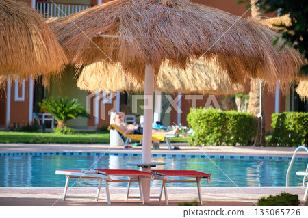 Empty deck chairs under straw shade umbrellas on swimming pool side in tropical resort. Summer vacations and getaway concept Empty deck chairs under straw shade umbrellas on swimming pool side in tropical resort. Summer vacations and getaway concept 135065726