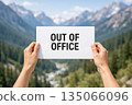 Two raised hands hold a white paper sign with Out of Office text with clear blue sky and mountain.. 135066096
