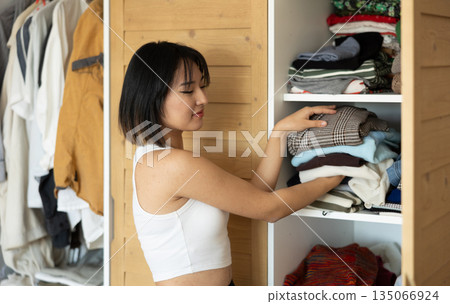 Happy young Asian girl putting some items of clothing in the wardrobe Happy young Asian girl putting some items of clothing in the wardrobe 135066924