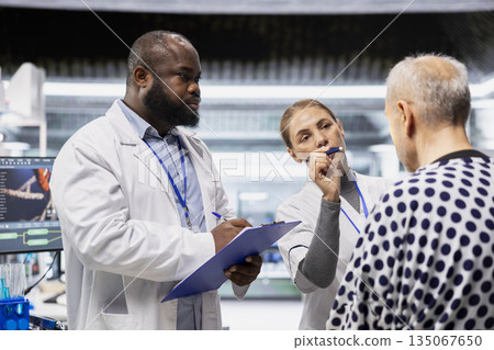 Two specialists participates in laboratory drug study monitoring a patient during treatment testing, recording dosage and side effects. Evaluating pharmacology data for healthcare progress. Two specialists participates in laboratory drug study monitoring a patient during treatment testing, recording dosage and side effects. Evaluating pharmacology data for healthcare progress. 135067650