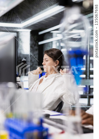Laboratory scientist relaxing back while using computer to process DNA patient data for clinical research. Lab specialist stretching sore muscles at PC desk, doing genetic analysis diagnostics Laboratory scientist relaxing back while using computer to process DNA patient data for clinical research. Lab specialist stretching sore muscles at PC desk, doing genetic analysis diagnostics 135067698