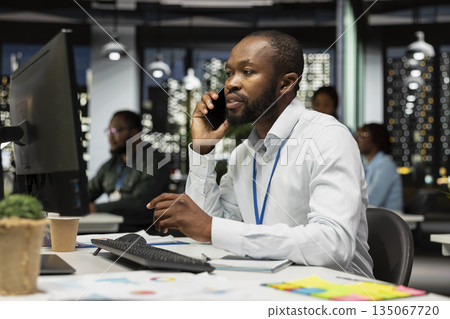 Business manager answers incoming phone call after hours at the office, using laptop to read briefing data and engage in a transparent dialogue. Exchanges ideas to strengthen relations. 135067720